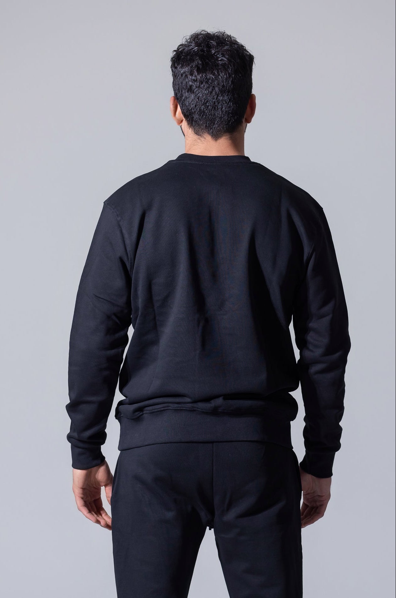 A man viewed from behind wearing a black crewneck sweatshirt and matching black sweatpants against a solid gray studio background.