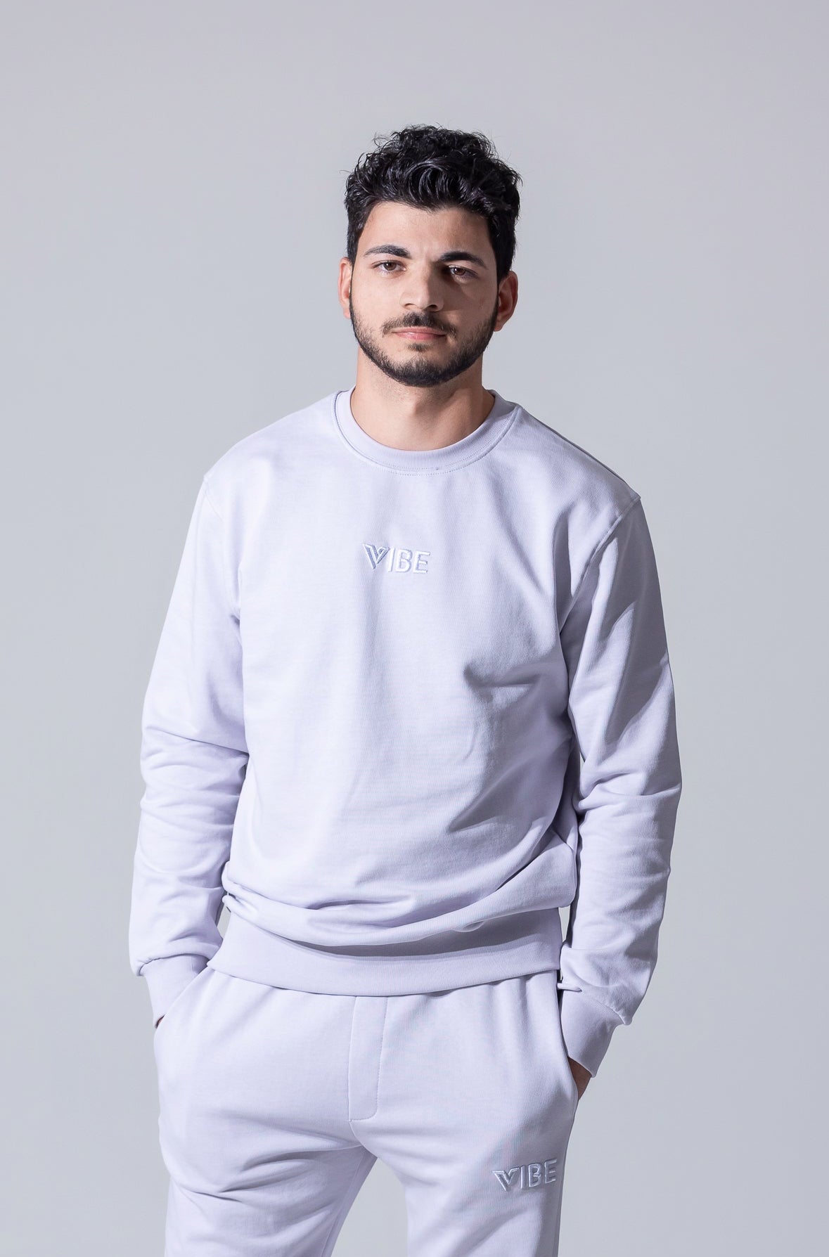A man wearing a light gray crewneck sweatshirt and matching sweatpants with a subtle 'VIBE' logo on the chest and thigh, photographed against a solid light gray background.