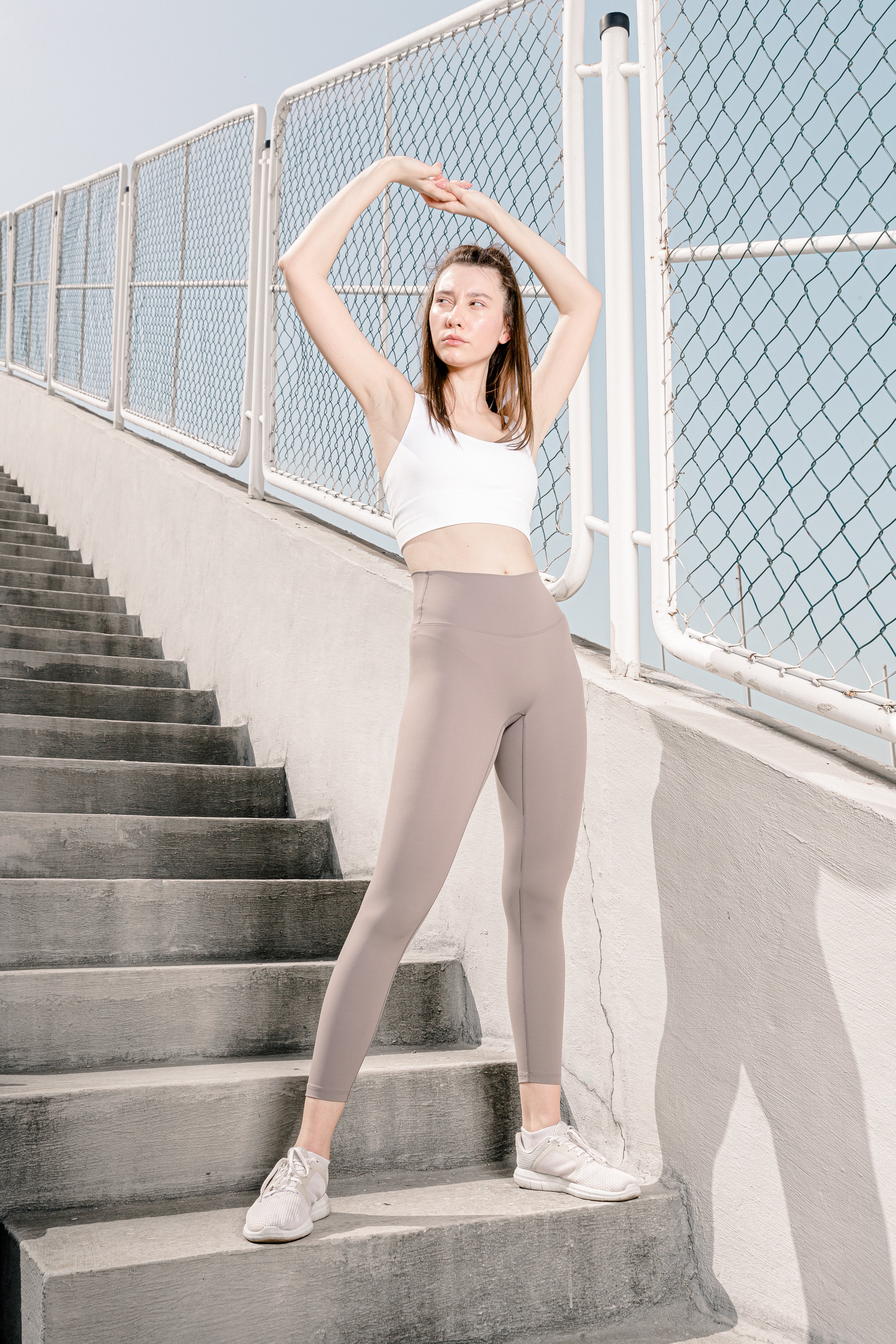 A person standing on a staircase wearing high-waisted beige leggings and a white sports top, with a chain-link fence and ocean in the background.