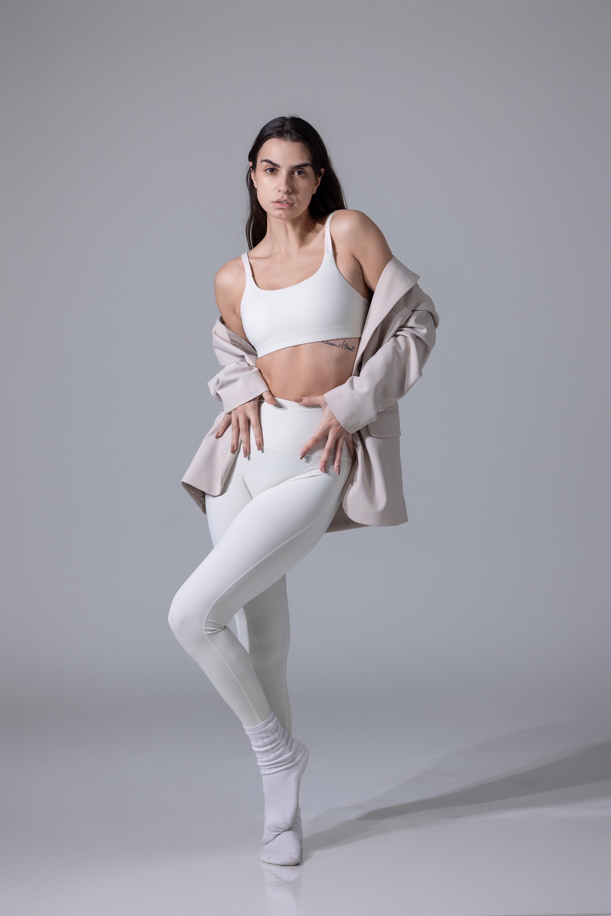 Woman in a white sports bra and leggings with a beige cardigan on a gray background