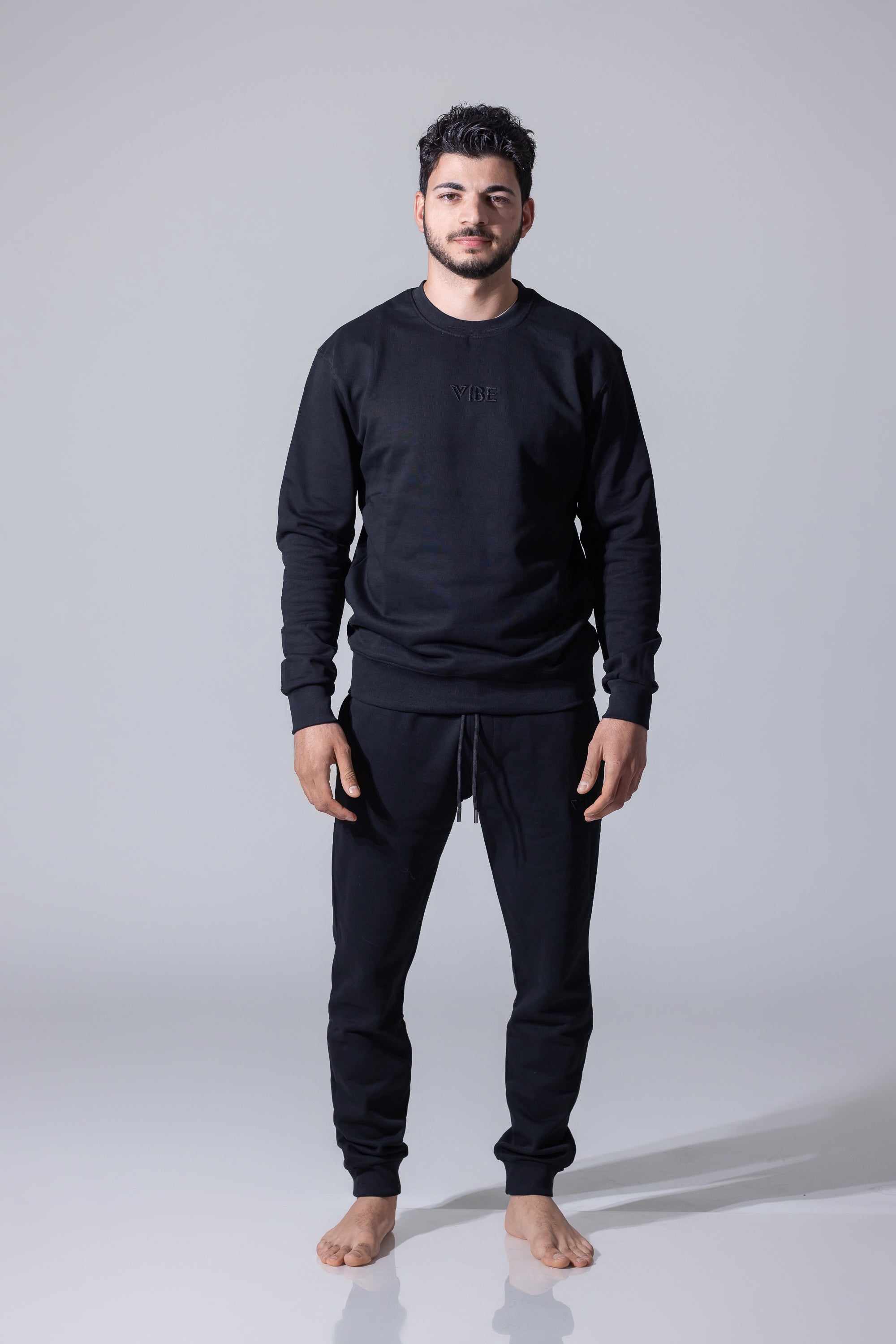A man wearing a matching black crewneck sweatshirt and jogger pants set, standing barefoot against a light gray studio background.