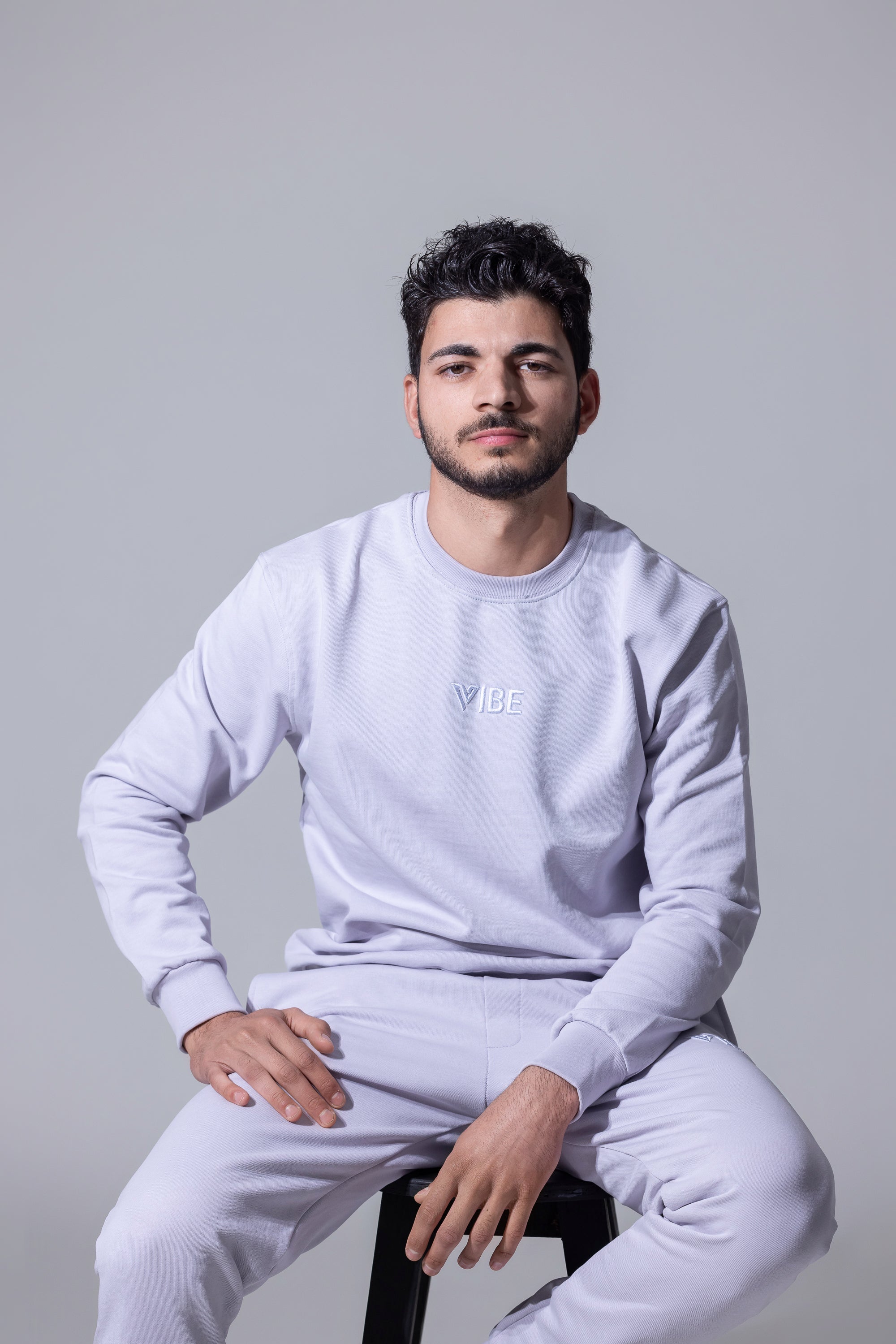 A man wearing a matching black crewneck sweatshirt and jogger pants set, sitting barefoot on a black stool against a light gray studio background.