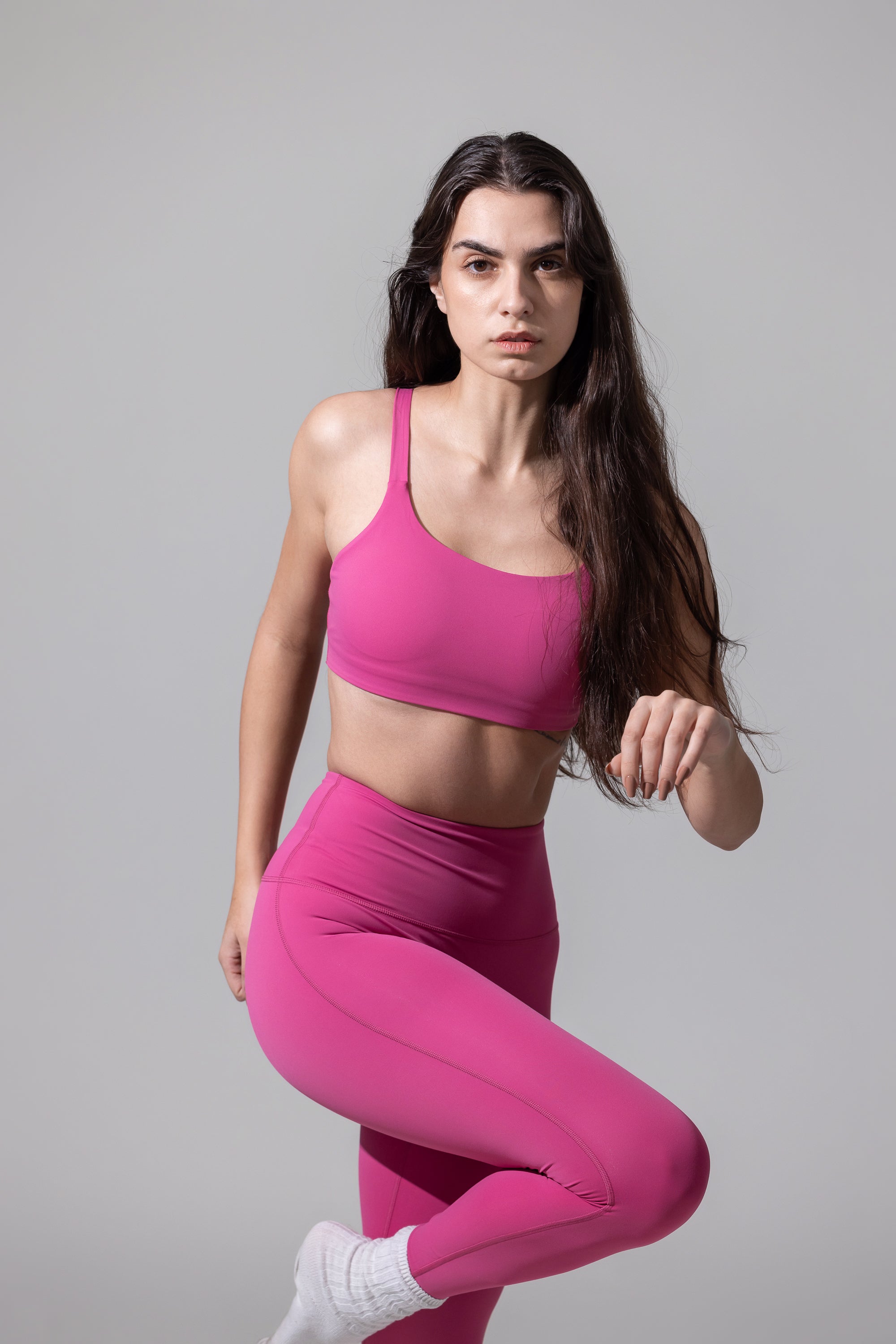 A woman wearing a pink sports bra and matching leggings, standing on one leg with her hand extended downwards. The leggings are solid pink, and the bra is also pink with a mesh design.