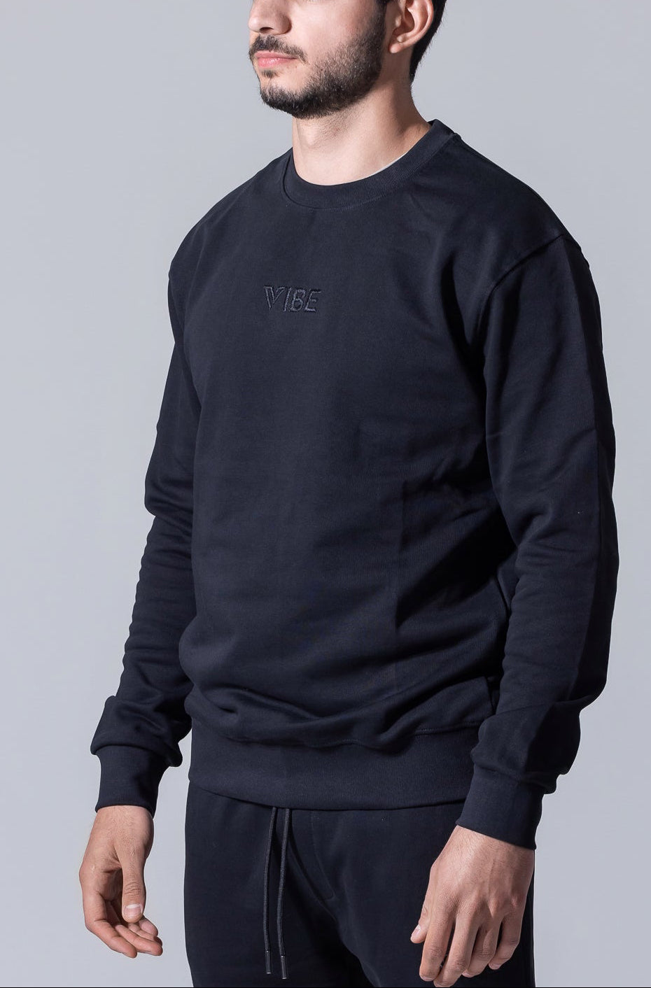 A man wearing a black crewneck sweatshirt with a subtle tonal embroidered logo on the chest, paired with matching dark pants, set against a plain gray studio background.