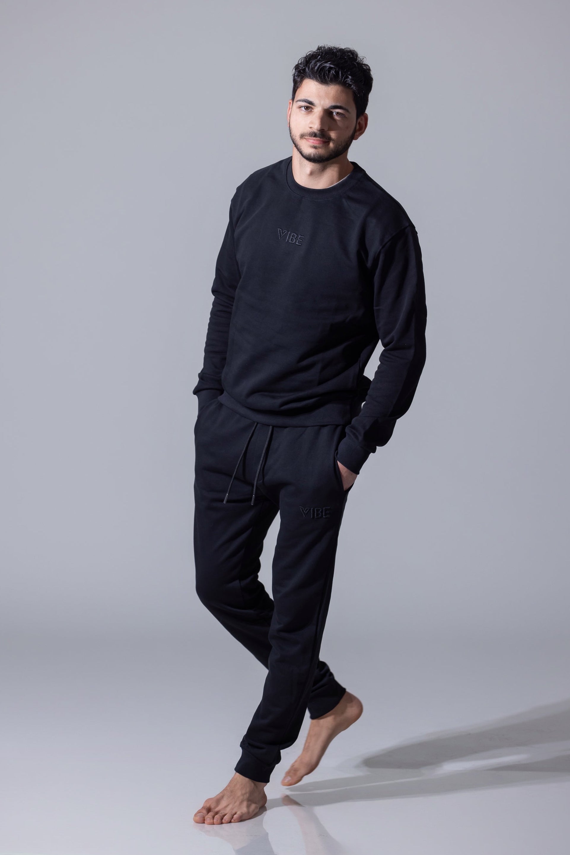 A male model standing on a white background, wearing black men Sweatpants with a neutral finish. He is also wearing a black long-sleeve shirt and black shoes.
