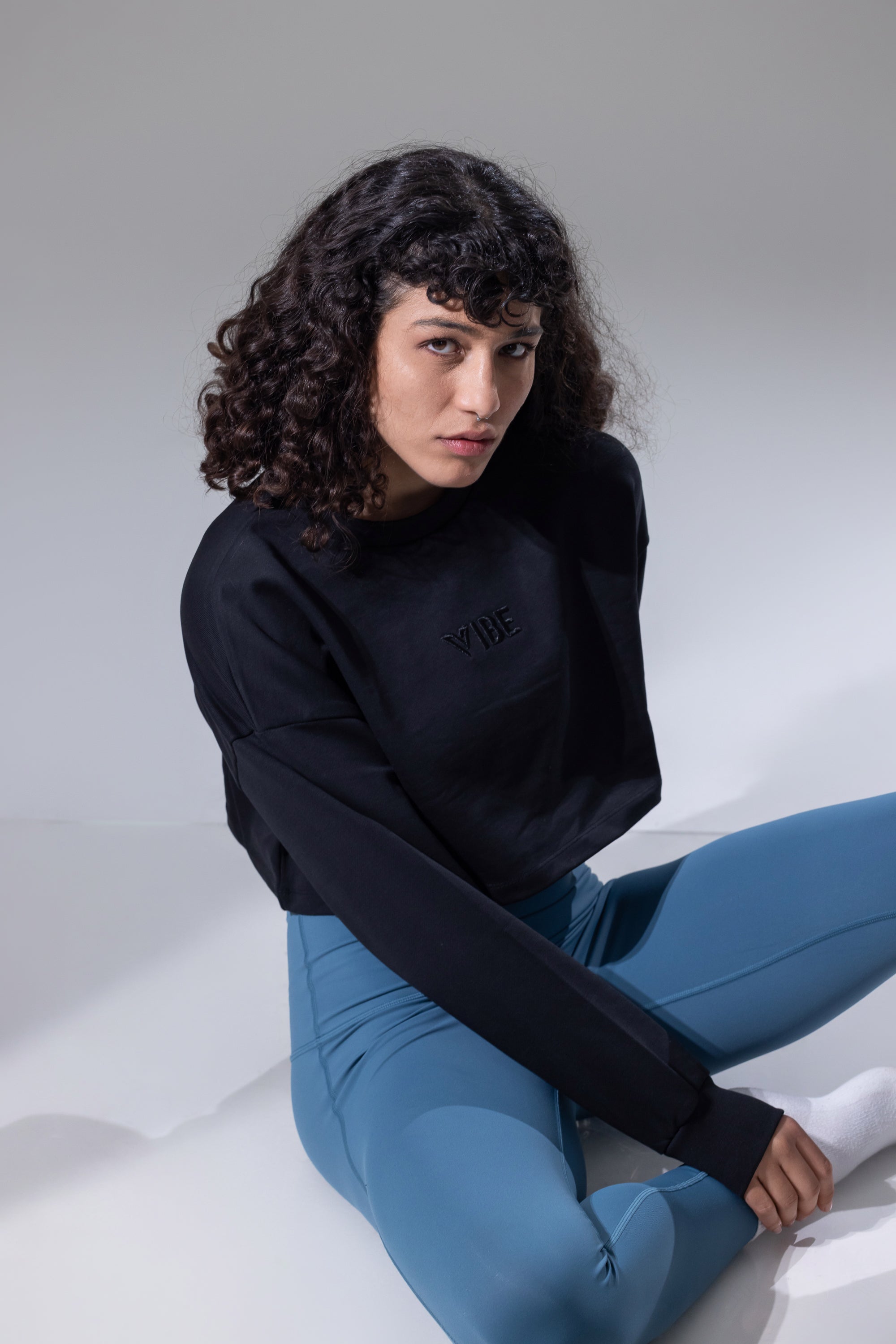 A woman wearing a black long-sleeve cropped sweatshirt with a tonal embroidered logo on the chest and bright blue leggings.