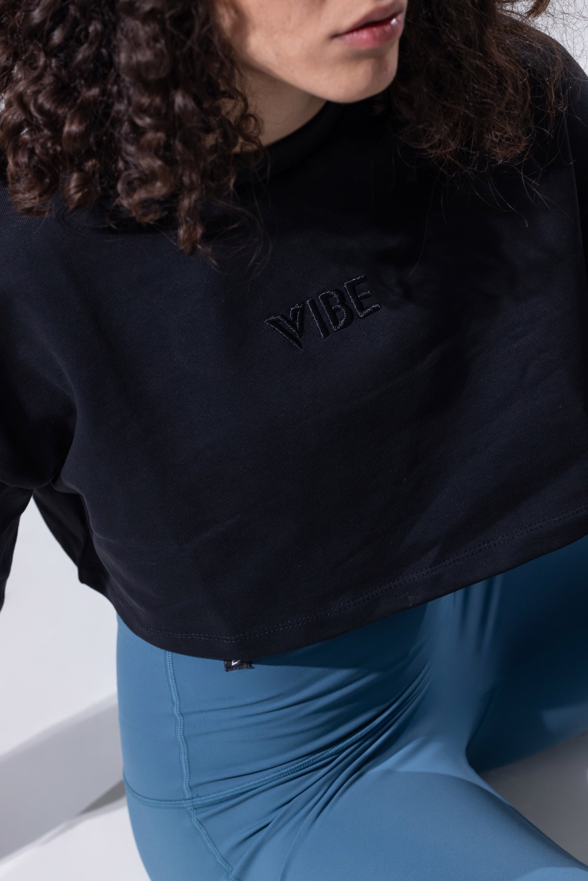 A close-up of a person wearing a black cropped sweatshirt with a tonal embroidered 'VIBE' logo on the chest, paired with blue leggings.