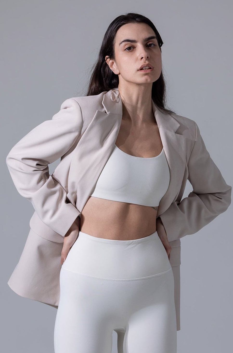 Woman wearing a light jacket, white sports bra, and leggings on a gray background