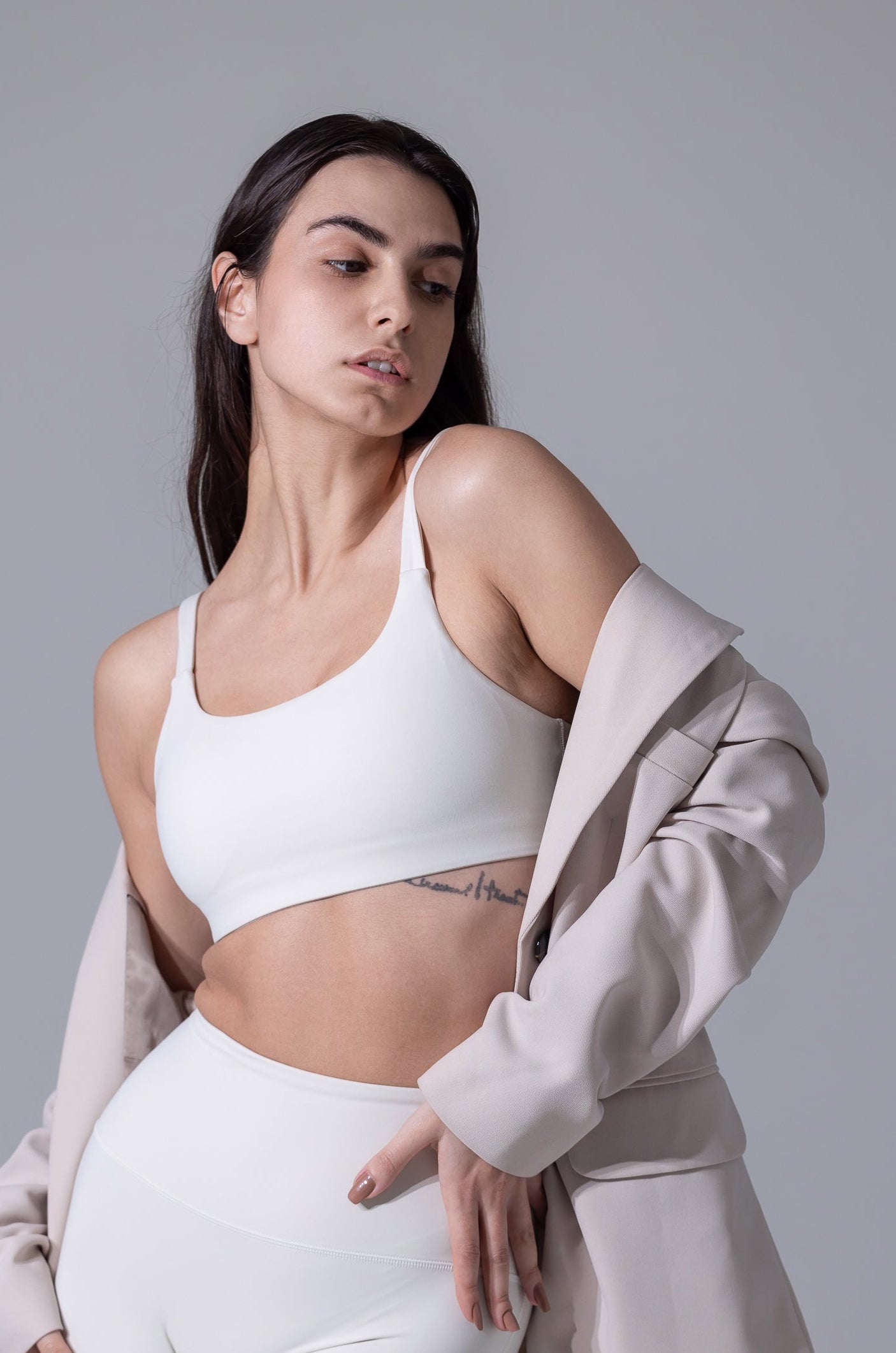 Woman wearing a white sports bra and leggings with a beige cardigan on a gray background