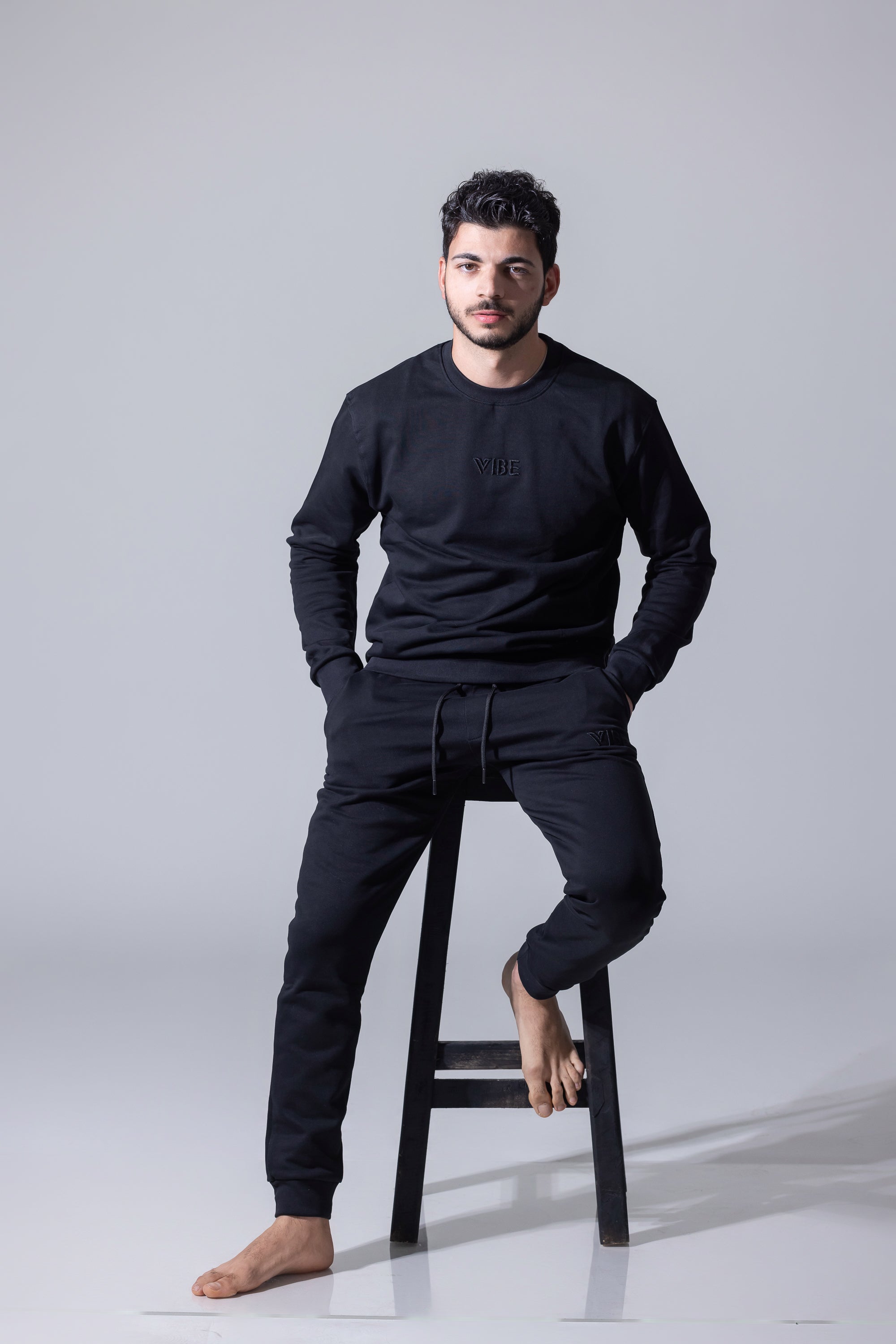 A man wearing a matching black crewneck sweatshirt and jogger pants set, sitting barefoot on a black stool against a light gray studio background.