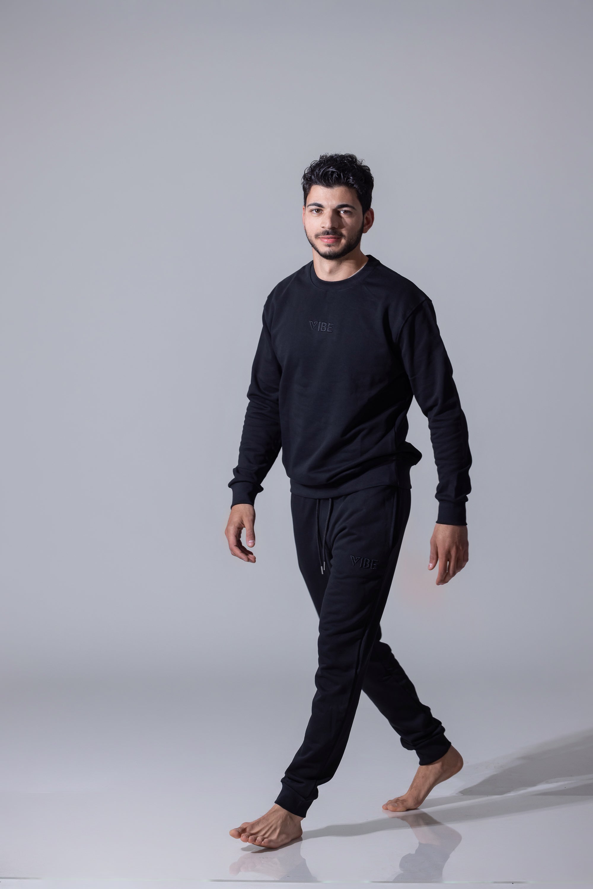 A man wearing a matching black long-sleeve sweatshirt and ribbed black pants, walking barefoot on a light gray studio background.