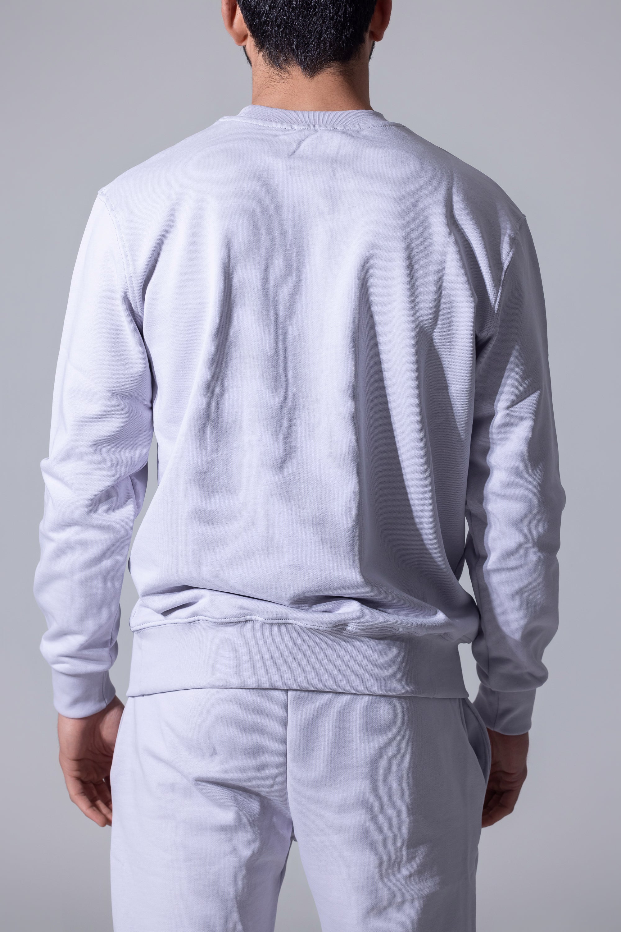 A man viewed from behind wearing a light gray crewneck sweatshirt and matching sweatpants against a neutral gray studio background.